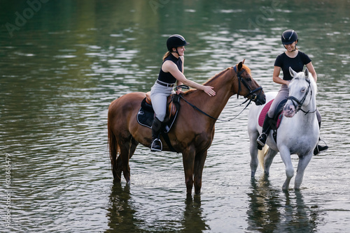 Riders, two young women riding beautiful horses down the calm river surrounded by the green grove © 24K-Production