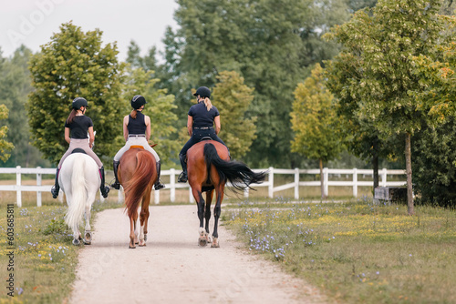 Rear view of three female riders riding horses side by side near white wood fencing, returning to the horse farm