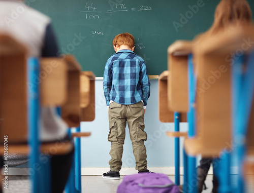 Rear view, boy student in detention with head on chalkboard and in classroom of school building. Anxiety or depressed for time out, math problems to solve and male kid with back to class. photo