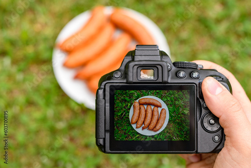 Kiełbasa grillowa na ekranie aparatu fotograficznego i leżąca na talerzu na trawie na świeżym powietrzu 