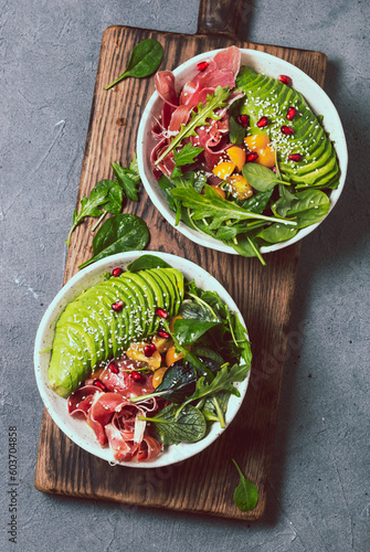 Low carbs bowl. Fresh salad with green spinach, rucola, avocado an ham serrano in white bowl, gray background, top view photo