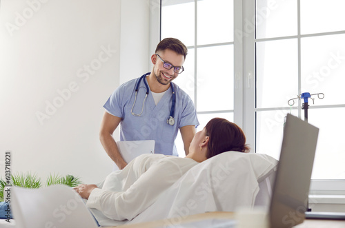 Friendly male nurse and female patient at clinic. Smiling young man in scrubs uniform sets intravenous IV line infusion system for overweight woman lying on medical bed in hospital room