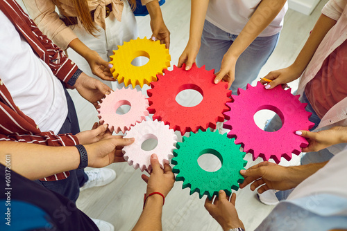 Team of young people standing in circle connecting colorful yellow, red, pink, green cogwheels. Cropped shot of hands holding different gears. Teamwork, business, education, success concept background