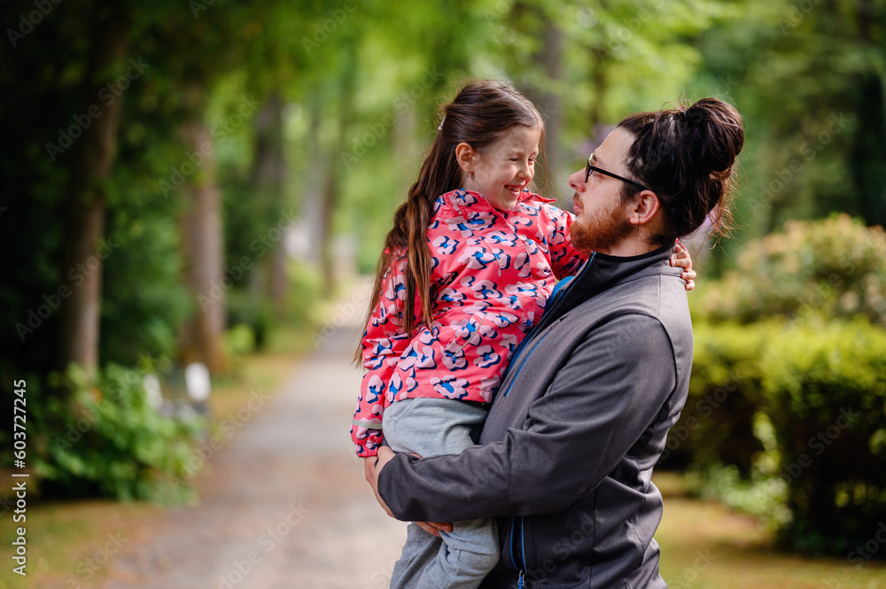 Fototapeta premium Young long hair bearded man in glasses standing with junior girl holding on hands on road in park