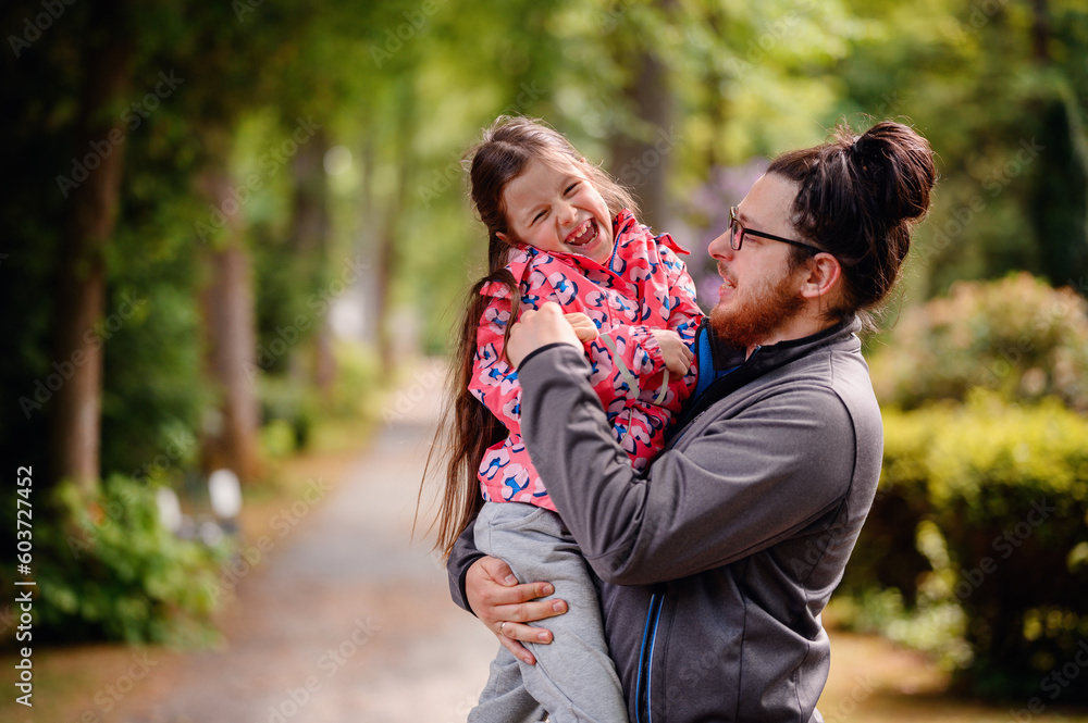 Young long hair bearded man in glasses standing with junior girl holding on hands on road in park Young long hair bearded man in glasses standing with junior girl holding on hands on road in park