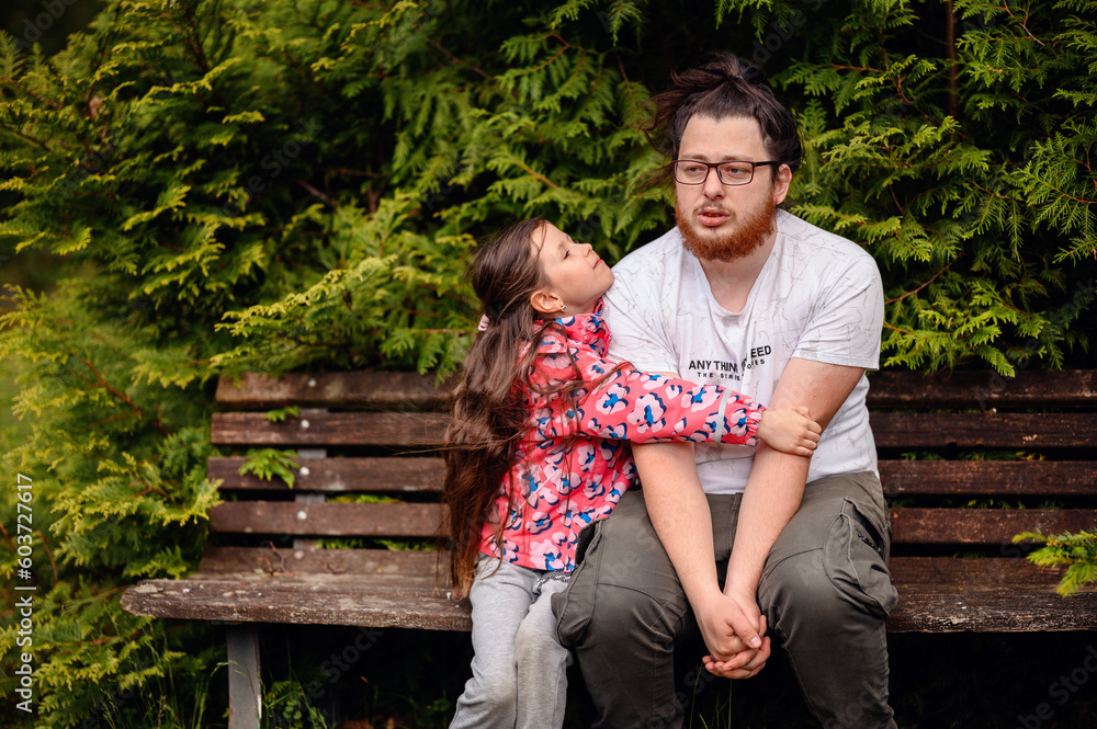 Young long haired man in glasses sits with junior girl on wooden bench Young long haired man in glasses sits with junior girl on wooden bench