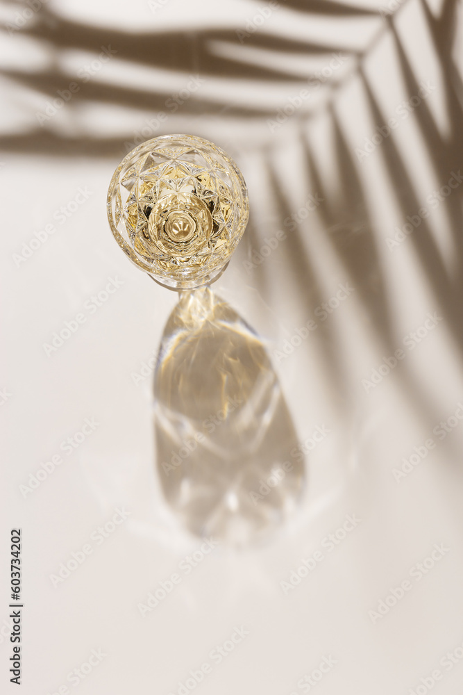 White wine in crystal transparent fashion glass with palm leaf shadow at sunlight, beige background and beautiful light, creative summer alcohol drink minimal concept, copyspace, top view