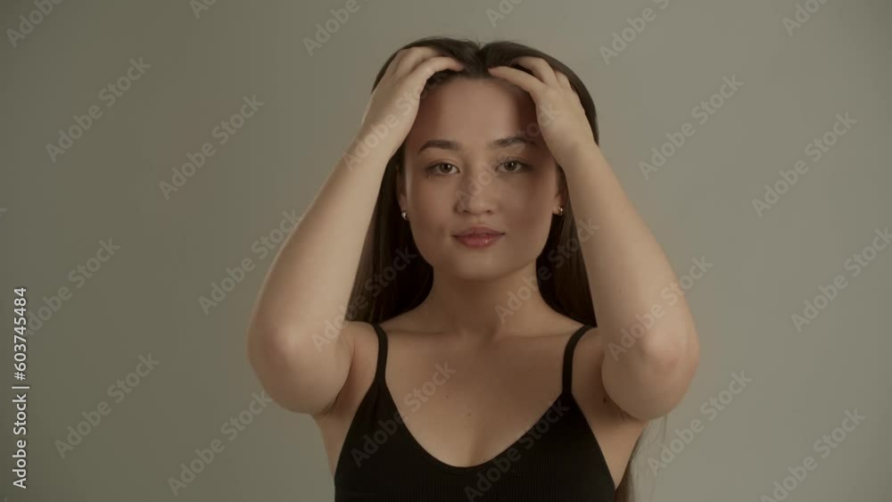 A young long-haired woman stands in front of the camera and fixes her hair. A young woman standing on a gray background gently strokes her face with her fingers. A young long-haired woman stands in front of the camera and fixes her hair. A young woman standing on a gray background gently strokes her face with her fingers.