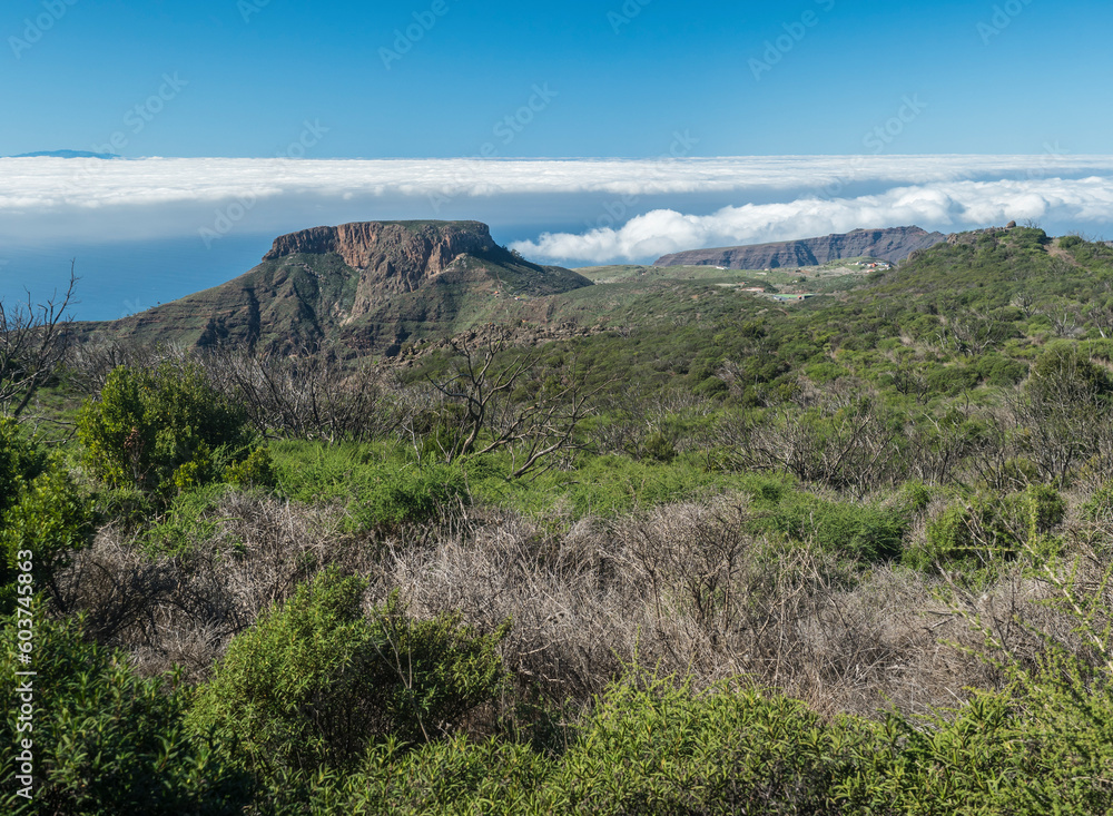 Scenic view on table mountain Fortaleza seen from peak of Alto de at Garajonay mountain. White clouds and El Hiero island above, blue sky. La Gomera, Canary Islands, Spain. Scenic view on table mountain Fortaleza seen from peak of Alto de at Garajonay mountain. White clouds and El Hiero island above, blue sky. La Gomera, Canary Islands, Spain.