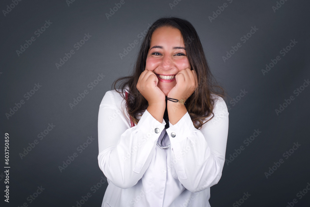 Portrait of young cute european doctor woman being overwhelmed with emotions, expressing excitement and happiness with closed eyes and hands near face while smiling broadly over gray background. Portrait of young cute european doctor woman being overwhelmed with emotions, expressing excitement and happiness with closed eyes and hands near face while smiling broadly over gray background.