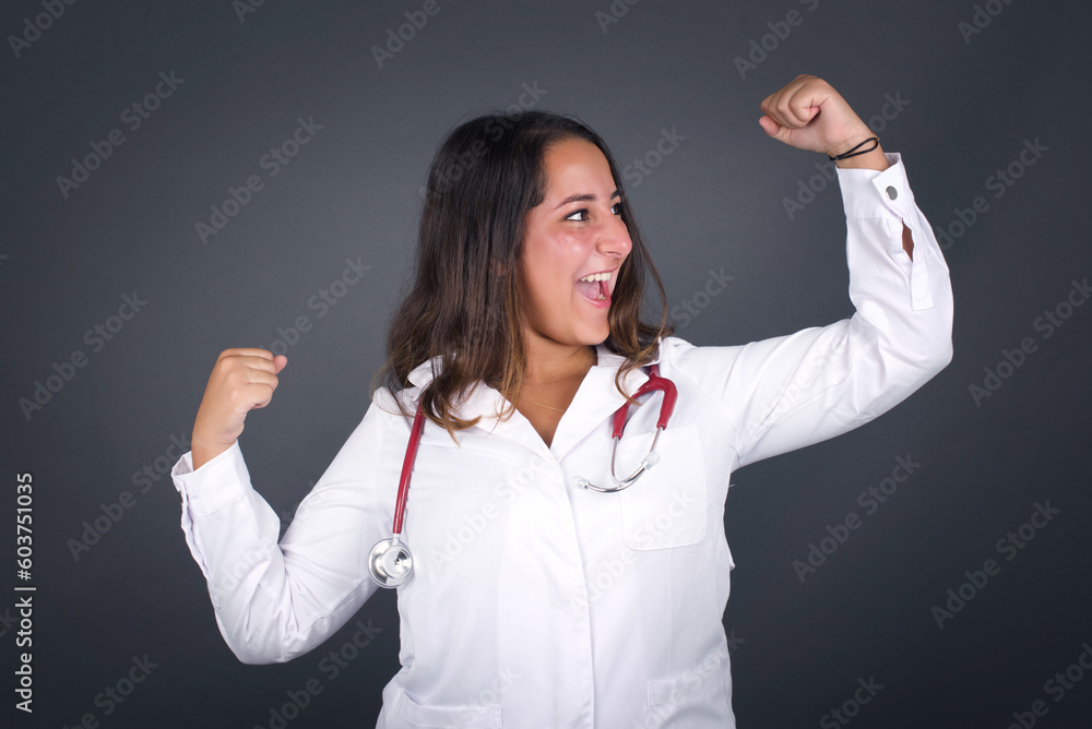 Young beautiful doctor woman wearing medical uniform over gray studio background celebrating a victory punching the air with her fists and a beaming toothy smile Young beautiful doctor woman wearing medical uniform over gray studio background celebrating a victory punching the air with her fists and a beaming toothy smile