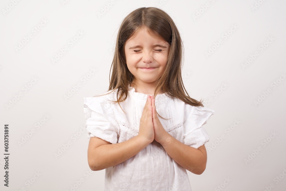 Cute charming little girl wearing white shirt over white background holding palms together, closing eyes and praying for peace. Cute charming little girl wearing white shirt over white background holding palms together, closing eyes and praying for peace.