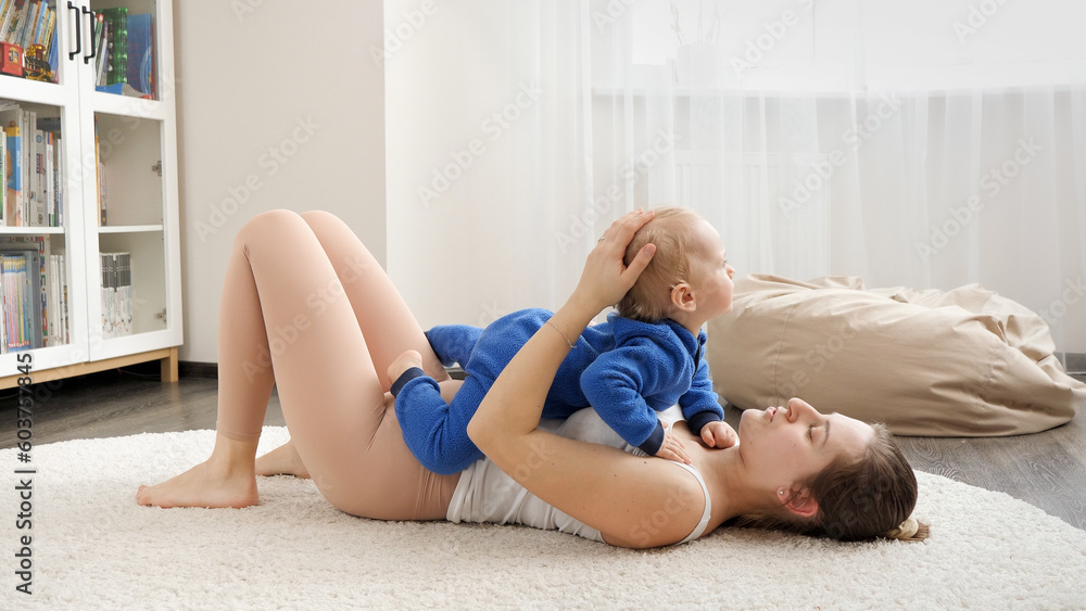 Obraz premium Young loving mother holding her baby son while doing fitness exercises on floor at home. Family healthcare, active lifestyle, parenting and child development