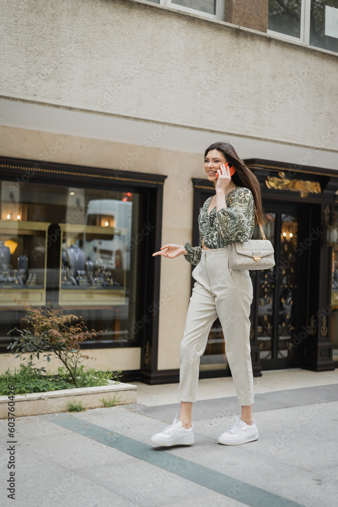 Fototapeta premium cheerful young woman with long hair and makeup smiling while talking on smartphone and walking in trendy outfit with handbag on chain strap near blurred jewelry store in Istanbul