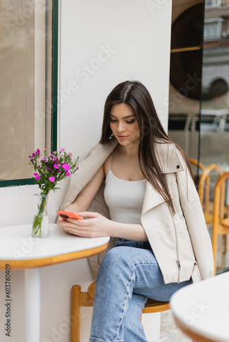 young woman with long hair sitting on chair near bistro table with flowers in vase and texting on smartphone while sitting in trendy clothes with leather jacket in cafe on terrace outdoors in Istanbul © LIGHTFIELD STUDIOS