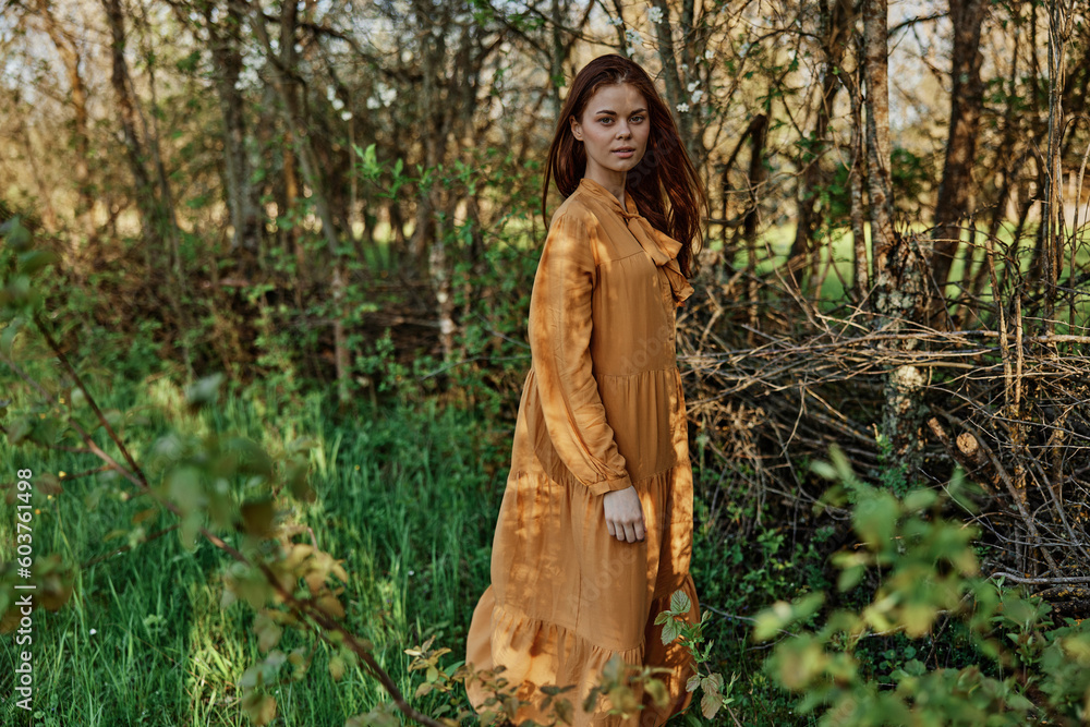 a beautiful woman walks in the shade near the trees, dressed in a long orange dress, enjoying the weather and the weekend, posing relaxed. The theme of privacy with nature a beautiful woman walks in the shade near the trees, dressed in a long orange dress, enjoying the weather and the weekend, posing relaxed. The theme of privacy with nature