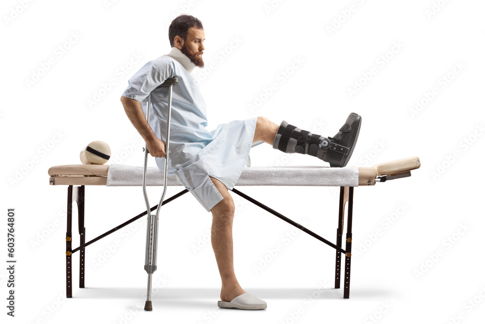 Man in a hospital gown sitting at a medical table with a broken leg and neck collar Man in a hospital gown sitting at a medical table with a broken leg and neck collar