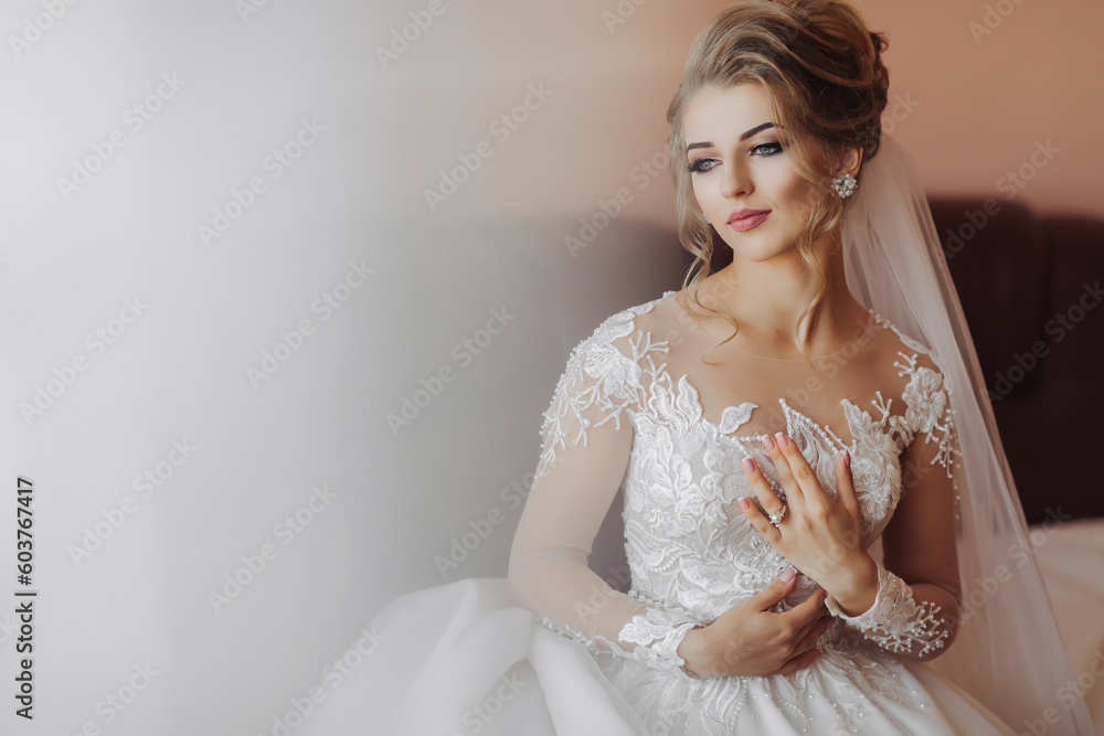 Blonde bride in long sleeve lace dress standing in her room, posing and putting on her engagement ring. Beautiful hair and makeup, open shoulders. Wedding portrait. Blonde bride in long sleeve lace dress standing in her room, posing and putting on her engagement ring. Beautiful hair and makeup, open shoulders. Wedding portrait.