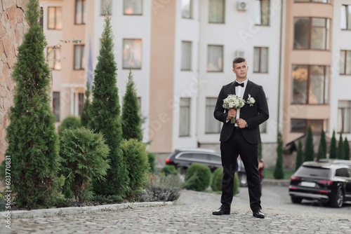 Portrait. A man in a white shirt, a black bow tie and a black suit holds a bouquet and goes to a meeting, behind him are black cars, new buildings and trees. A stylish watch. Men's style. Business © Vasil