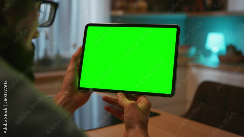 Shot of unrecognizable Caucasian man holding black digital tablet with green screen. Male hands using device with chroma key while sitting at table on blurred background of kitchen. Indoors Shot of unrecognizable Caucasian man holding black digital tablet with green screen. Male hands using device with chroma key while sitting at table on blurred background of kitchen. Indoors
