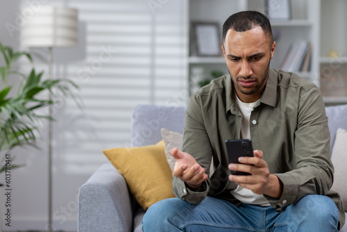 Fotografie Shocked young african american man sitting on sofa at home with phone in hands and looking worriedly at screen