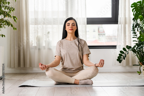 Young woman meditating sitting on a yoga mat near a big window photo