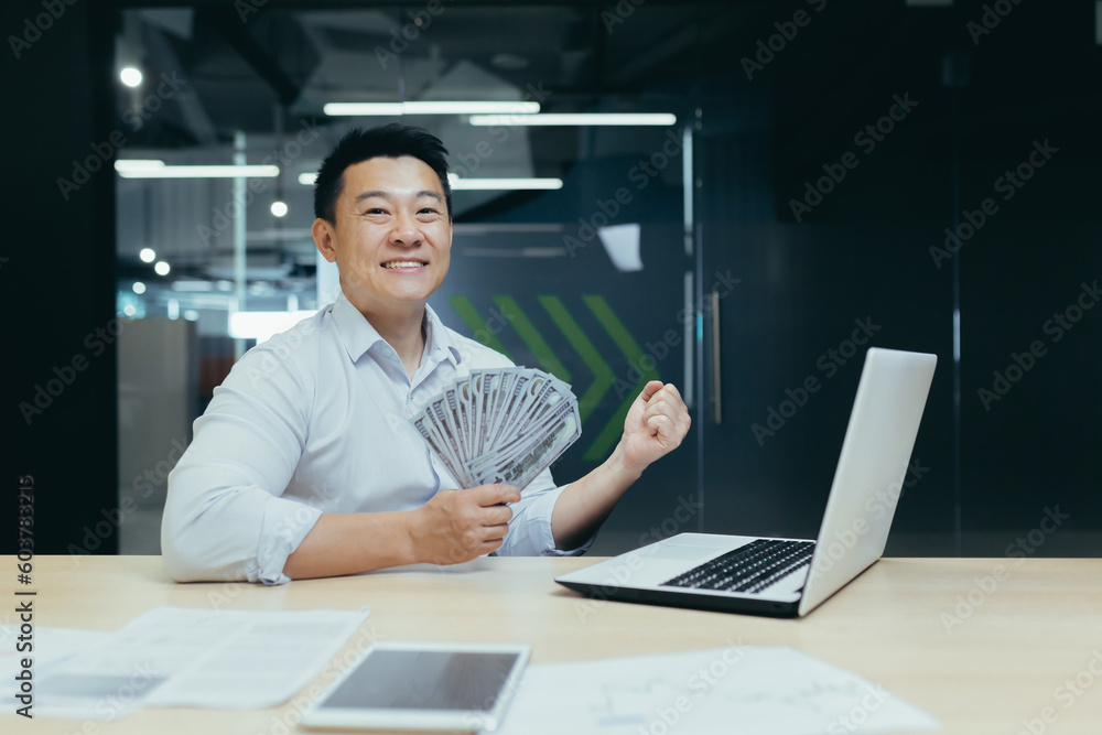 Portrait of a happy Asian man working in the office on a laptop, holding a fan of cash, dollar bills in his hand. shows a victory gesture with his hand to the camera, celebrates. Portrait of a happy Asian man working in the office on a laptop, holding a fan of cash, dollar bills in his hand. shows a victory gesture with his hand to the camera, celebrates.