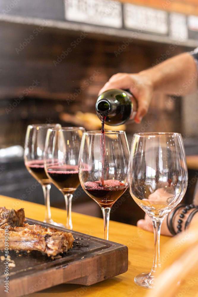 Fototapeta premium A white man hand serving a red wine bottle in four wine glasses in a row next to some gaucho traditional meat on a rustic wooden board in a familiar home or restaurant for lunch or dinner event