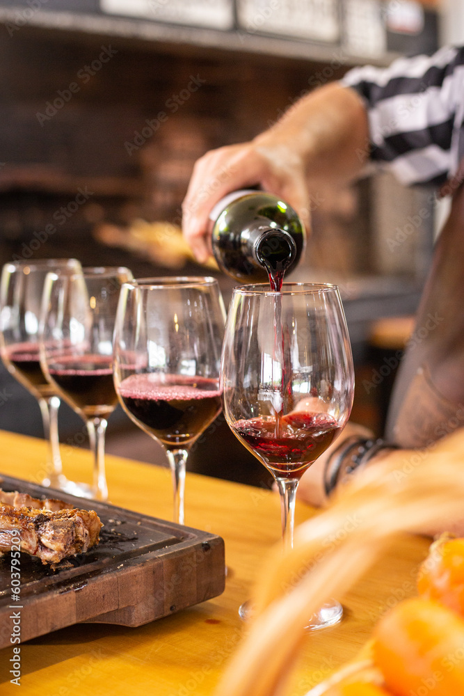 A white man serving the last of four glasses of red wine in a row next to a gaucho traditional barbecue on a rustic wooden board in a home or restaurant to have a nice lunch or dinner with some guests