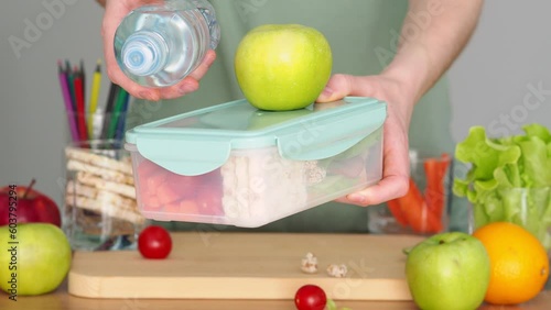 A woman holds out a lunchbox with food and water. Plastic lunch box and water bottle in female hands. Giving your child food