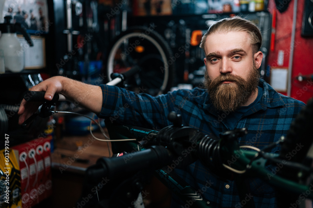 Closeup portrait of friendly cycling repairman standing behind bicycle in repair workshop with dark interior, looking at camera. Concept of professional repair and maintenance of bicycle transport.