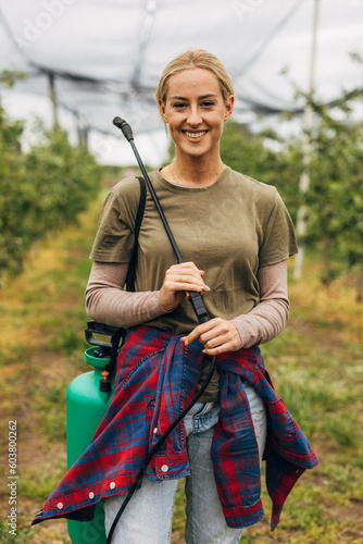 A pretty young woman holding a sprayer for plant protection stands in a fruit farm.