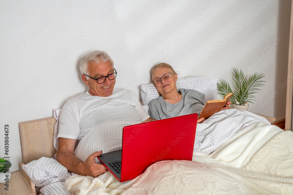 Senior couple spending leisure time in bed. A woman reads a book, a man works with a laptop while lying in the bedroom Senior couple spending leisure time in bed. A woman reads a book, a man works with a laptop while lying in the bedroom