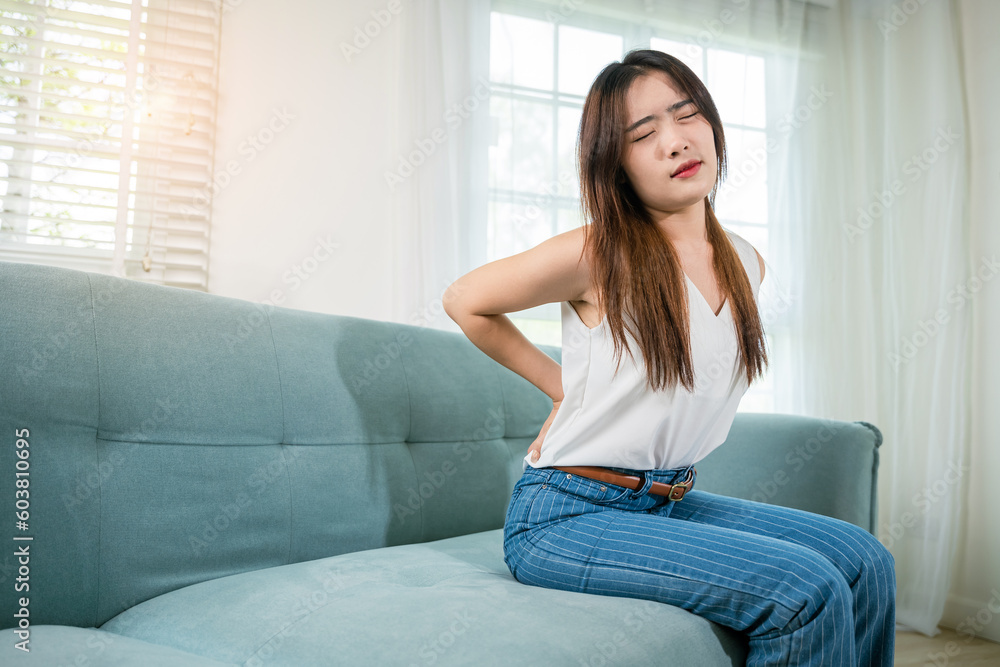 Asian woman holding her lower back feeling painful waist hurt massage in living room, beautiful young female touching back pain sitting in sofa she suffering from backache at home, healthcare medical Asian woman holding her lower back feeling painful waist hurt massage in living room, beautiful young female touching back pain sitting in sofa she suffering from backache at home, healthcare medical