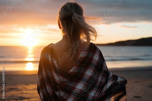 Blond haired girl covered with a cloth plaid blanket standing peacefully at the beach and looking into the ocean , generative AI