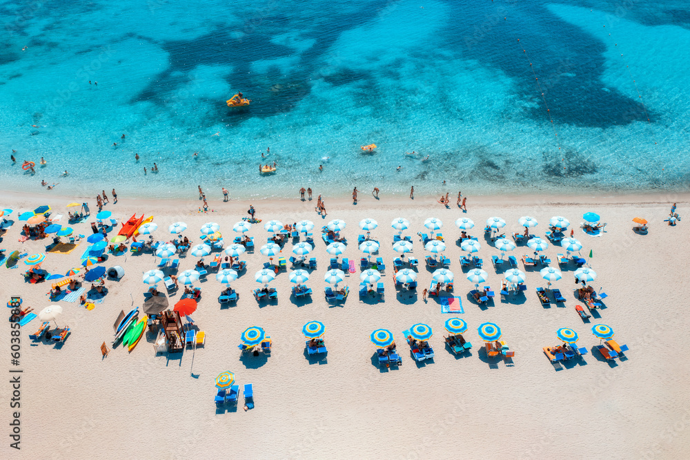 Aerial view of colorful umbrellas on sandy beach, swimming people in blue sea at summer sunny day. Sardinia, Italy. Tropical seascape. Travel and vacation. Top drone view of ocean with azure water