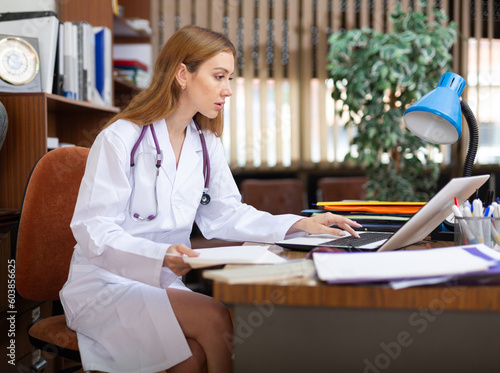 Young woman internist studies the patient's outpatient card, typing the treatment appointment on the computer, sitting at the ..workplace in the resident's office in the clinic