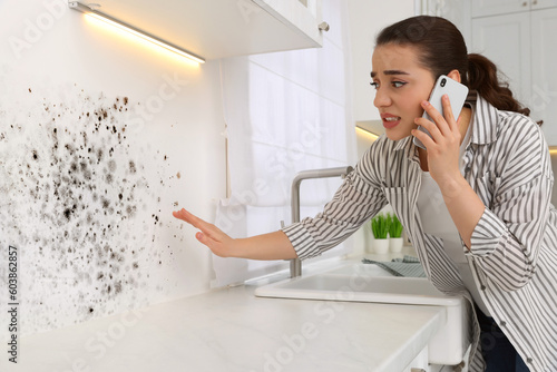 Mold removal service. Woman talking on phone and looking at affected wall in kitchen
