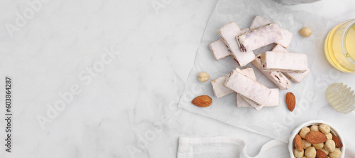 Pieces of delicious nutty nougat and ingredients on white marble table, flat lay. Banner design with space for text
