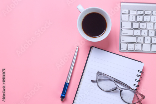 Feminine desk workspace with keyboard, notebook, glasses, pen and coffee cup on pink background. Flat lay with copy space