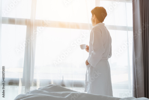 Tourist Asian man hold coffee cup in bathrobe towel looking at beautiful scene view on window at hotel room. Traveler Asian man feels relax in travel on holiday trip. Tourist relax in hotel concept.