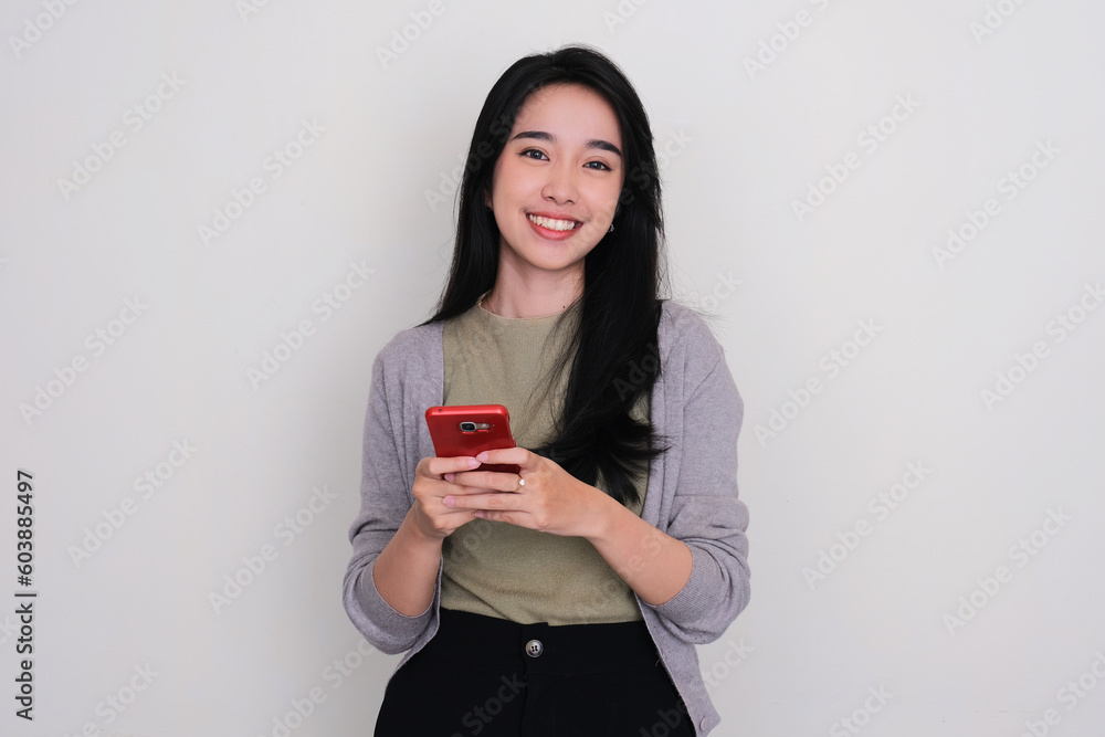 Naklejka premium Young Asian woman smiling at the camera while holding a mobile phone
