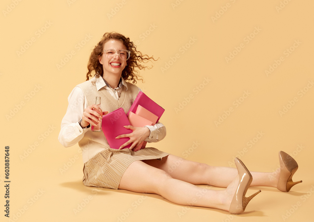 Portrait of pretty, curly, young girl sitting on floor in formal clothes, holding many books and drinking water over studio background. Concept of human emotions, youth culture, education, lifestyle Portrait of pretty, curly, young girl sitting on floor in formal clothes, holding many books and drinking water over studio background. Concept of human emotions, youth culture, education, lifestyle