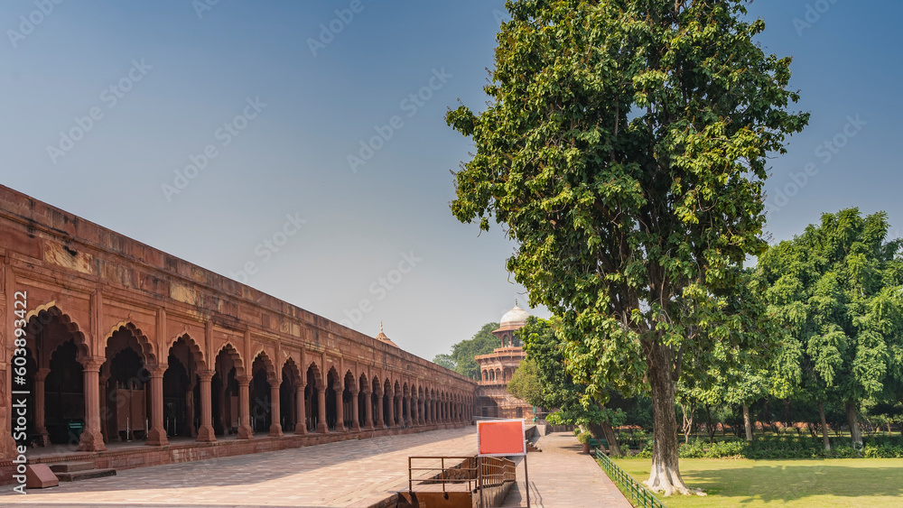 Fototapeta premium The ancient gallery is built around the perimeter of the Taj Mahal complex. Colonnade of red sandstone with arches. A tower with a dome in the distance. In the garden on the lawn - green grass, trees