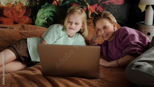 Caucasian woman and her preteen daughter relaxing together on bed at home watching series online on laptop photo