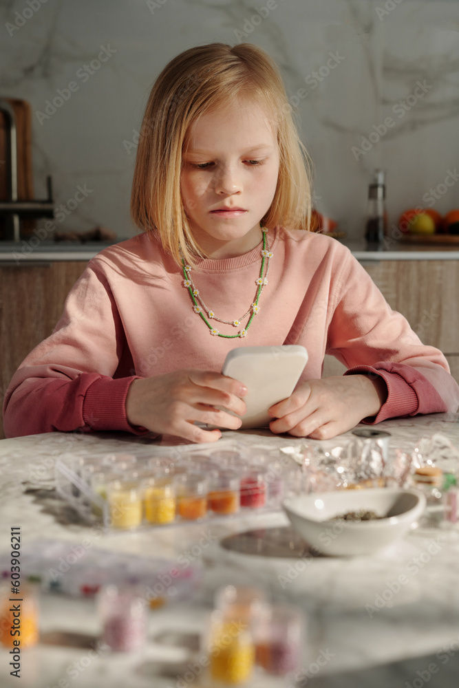 Vertical medium shot of preteen Caucasian girl sitting at table at home searching bead accessories design pictures for inspiration in Internet on smartphone Vertical medium shot of preteen Caucasian girl sitting at table at home searching bead accessories design pictures for inspiration in Internet on smartphone