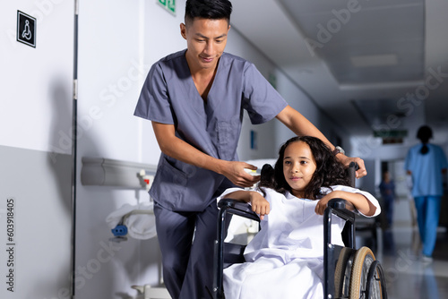 Smiling asian male doctor pushing biracial girl patient in wheelchair in corridor at hospital photo
