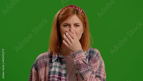 Disgusting odour. Discontent woman frowning face, smells something awful, feeling aversion, pink studio background. Woman closing nose bacause of a bad smell. Girl wear green knit sweater and hat. photo