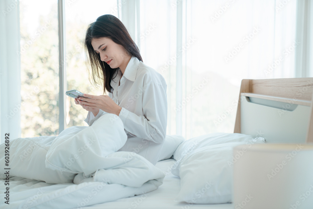 Photo of young happy woman in pajama and holding phone the phone to talk video call Online with friends while sitting on bed. Photo of young happy woman in pajama and holding phone the phone to talk video call Online with friends while sitting on bed.