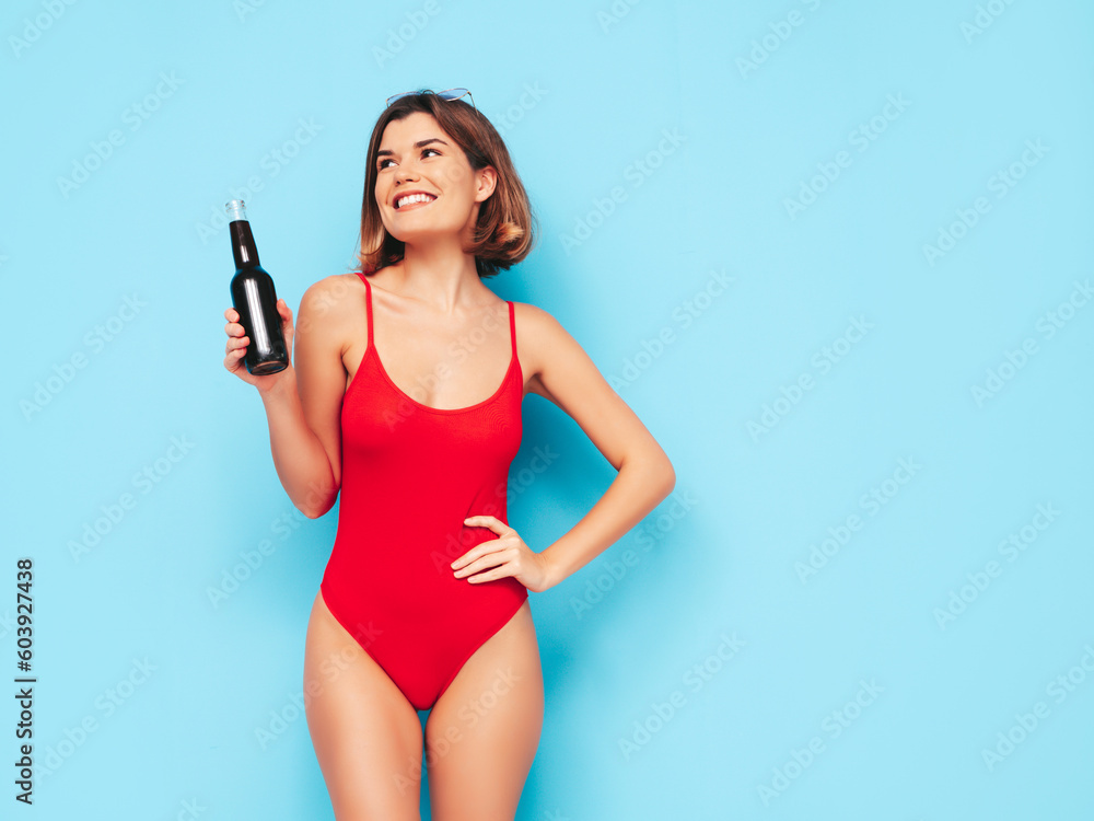 Young model in summer red swimwear bathing suit. Sexy carefree woman having fun and going crazy. Female posing near blue wall in studio. Drinking fresh cocktail smoozy drink from glass bottle Young model in summer red swimwear bathing suit. Sexy carefree woman having fun and going crazy. Female posing near blue wall in studio. Drinking fresh cocktail smoozy drink from glass bottle